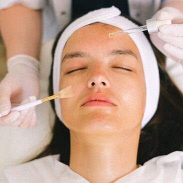 Top view skilled female cosmetologist in uniform and gloves applying essential oil on young woman face skin